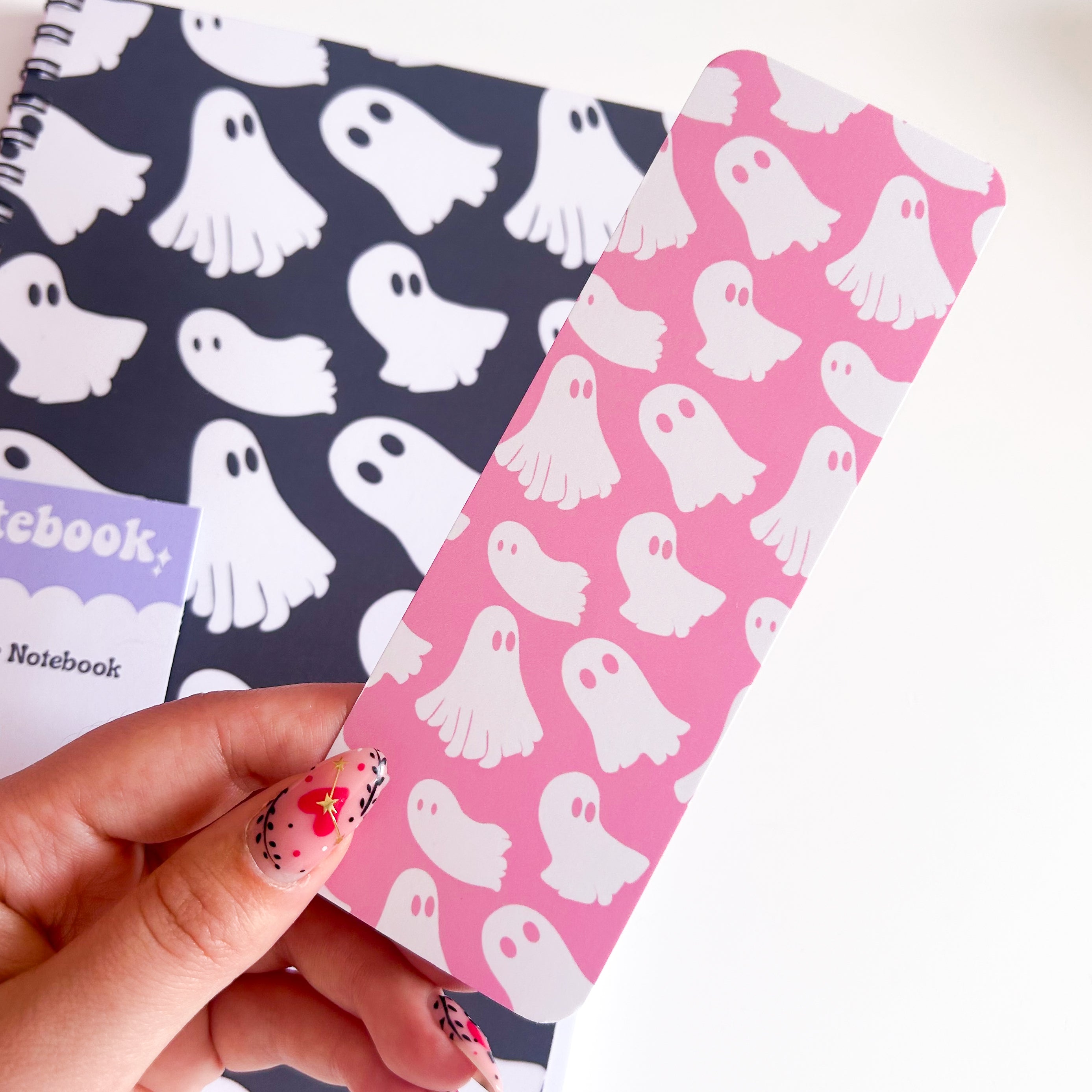Pink notebook with ghost pattern held by a hand, with a black notebook with ghost pattern in the background.