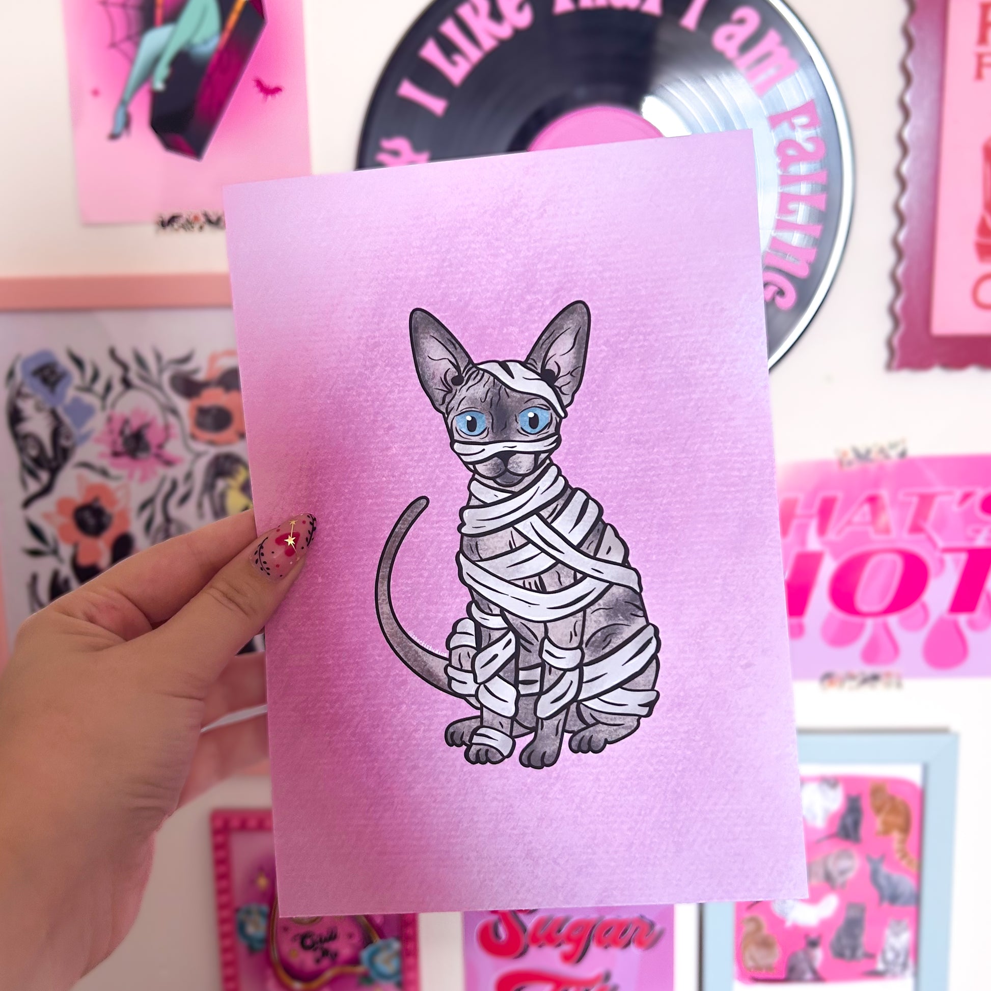 Hand holding a pink Sphynx cat illustration on a colorful background