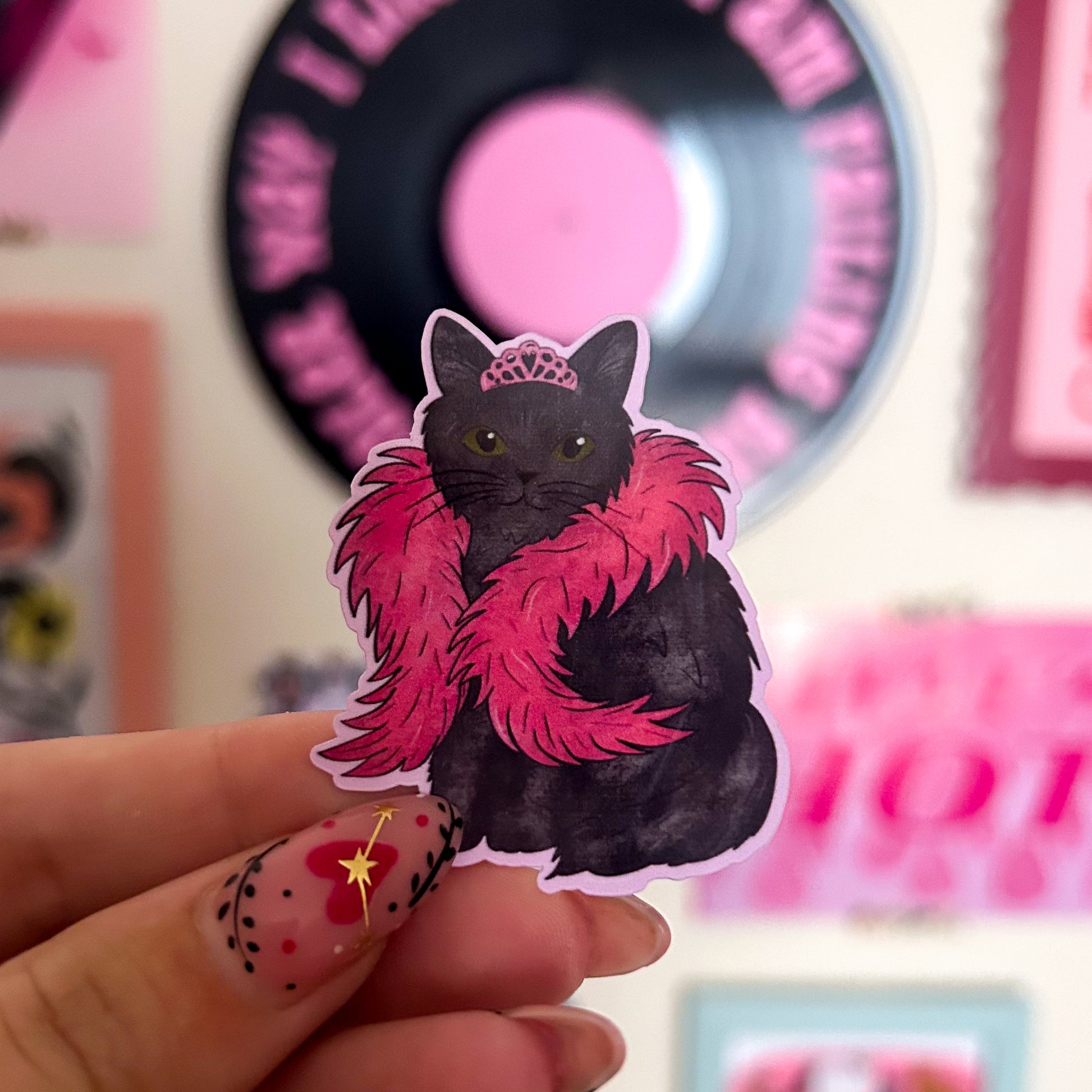 Sticker of a black cat with a pink boa and  pink tiara held by a hand against a blurred background. 