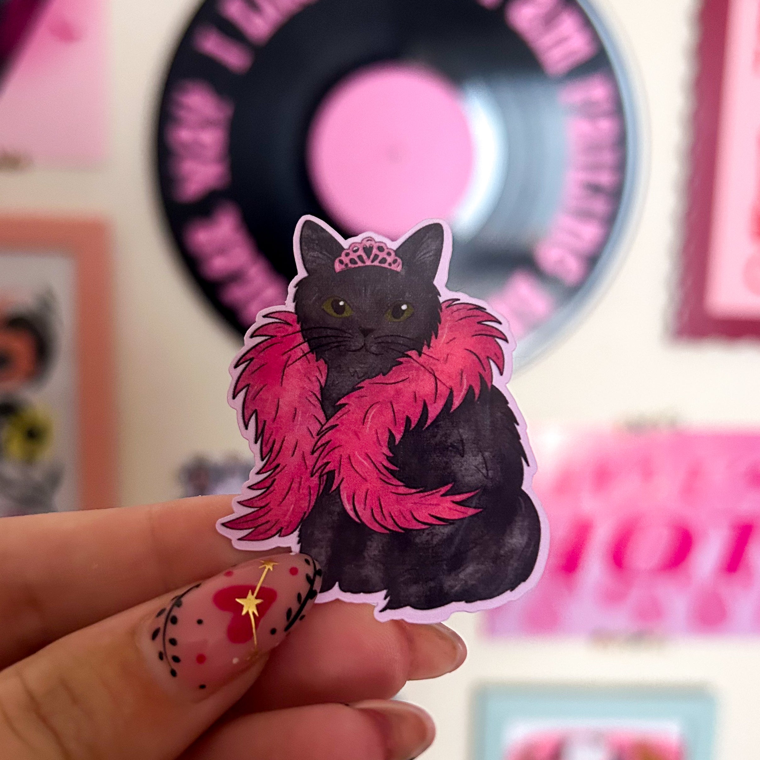 Sticker of a black cat with a pink boa and  pink tiara held by a hand against a blurred background. 
