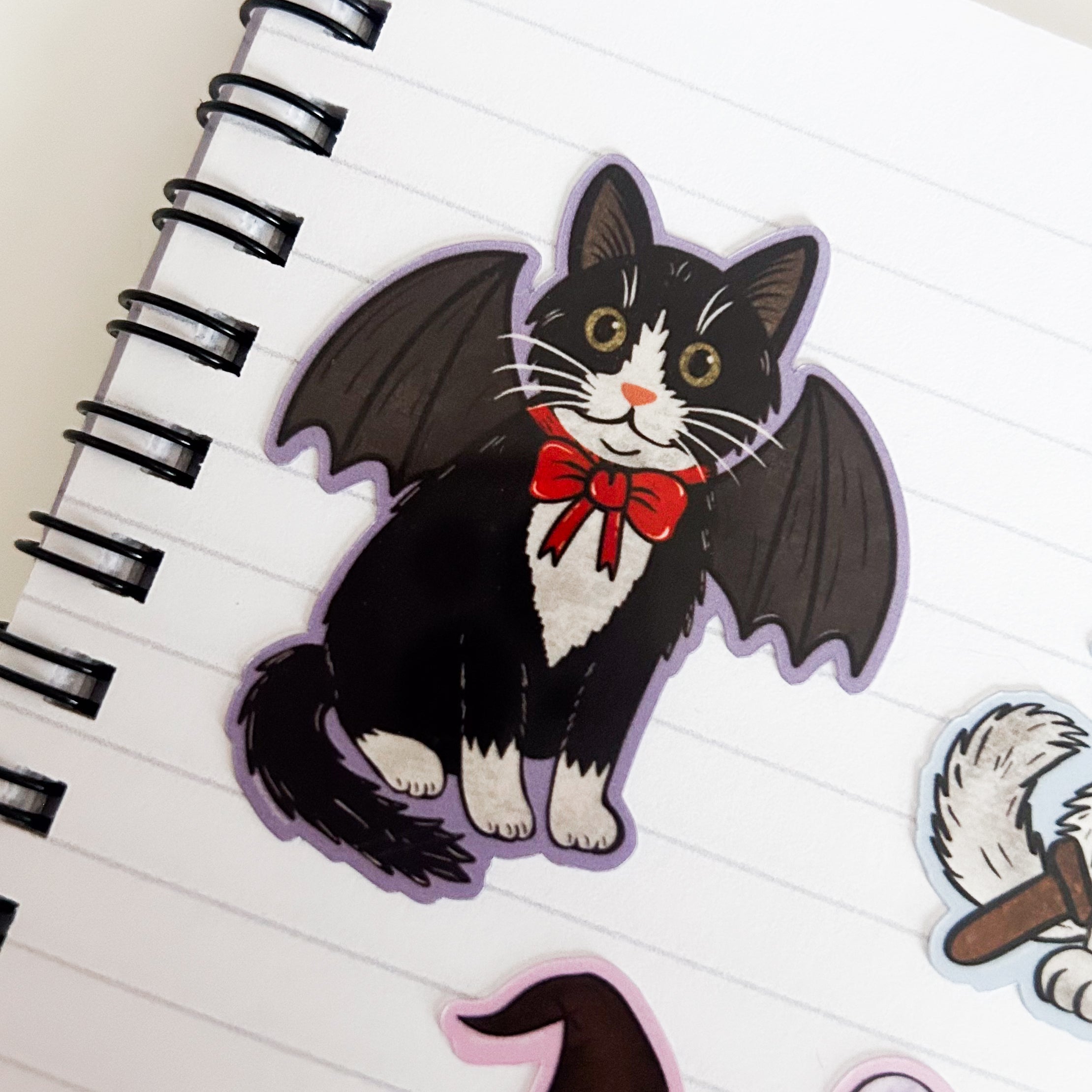 Sticker of a black and white tuxedo cat in a bat costume with bat wings and a red bow on white paper