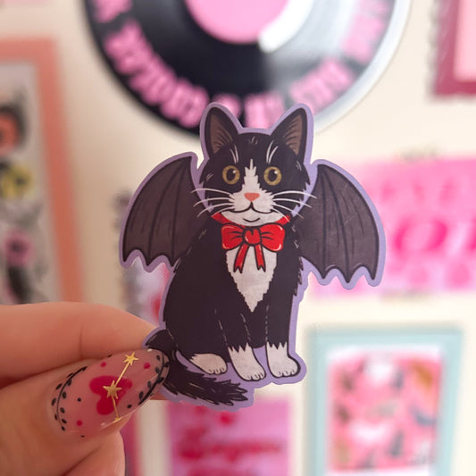 Sticker of a black and white tuxedo cat in a bat costume with bat wings and a red bow held by a hand against a colourful background