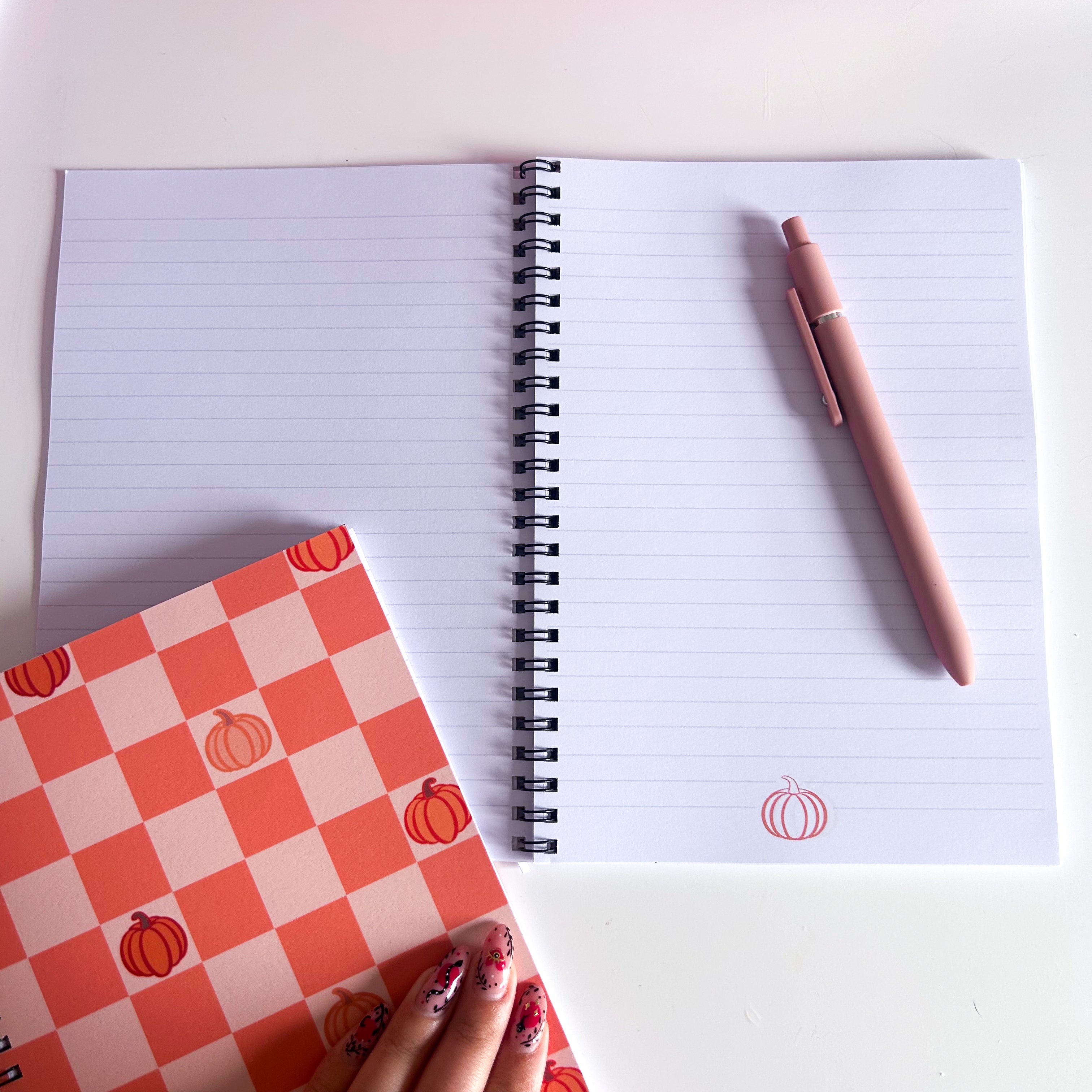 Open notebook with lined paper and an orange pumpkin illustration with a pink pen on a white surface, next to a small notebook with pumpkin pattern
