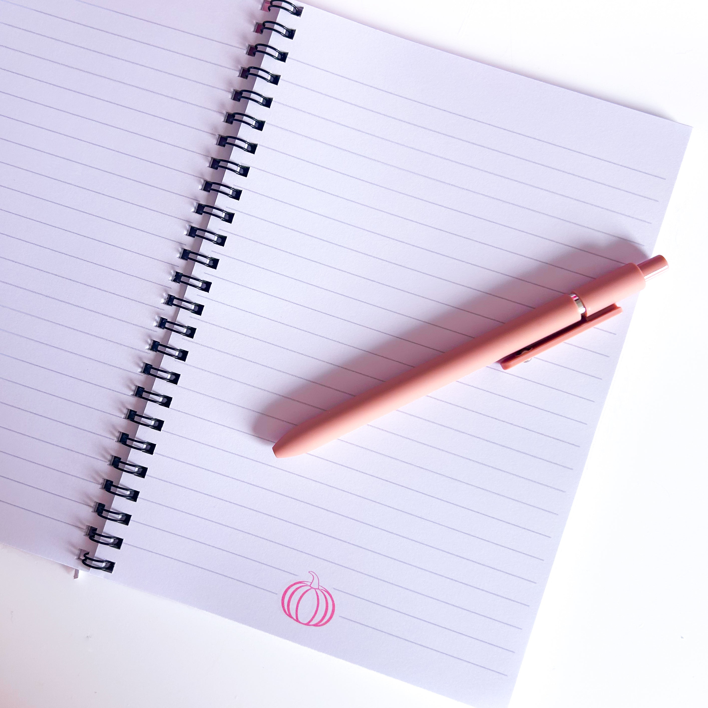 Open notebook with lined pages and a pink pumpkin illustration with a pink pen on a white background