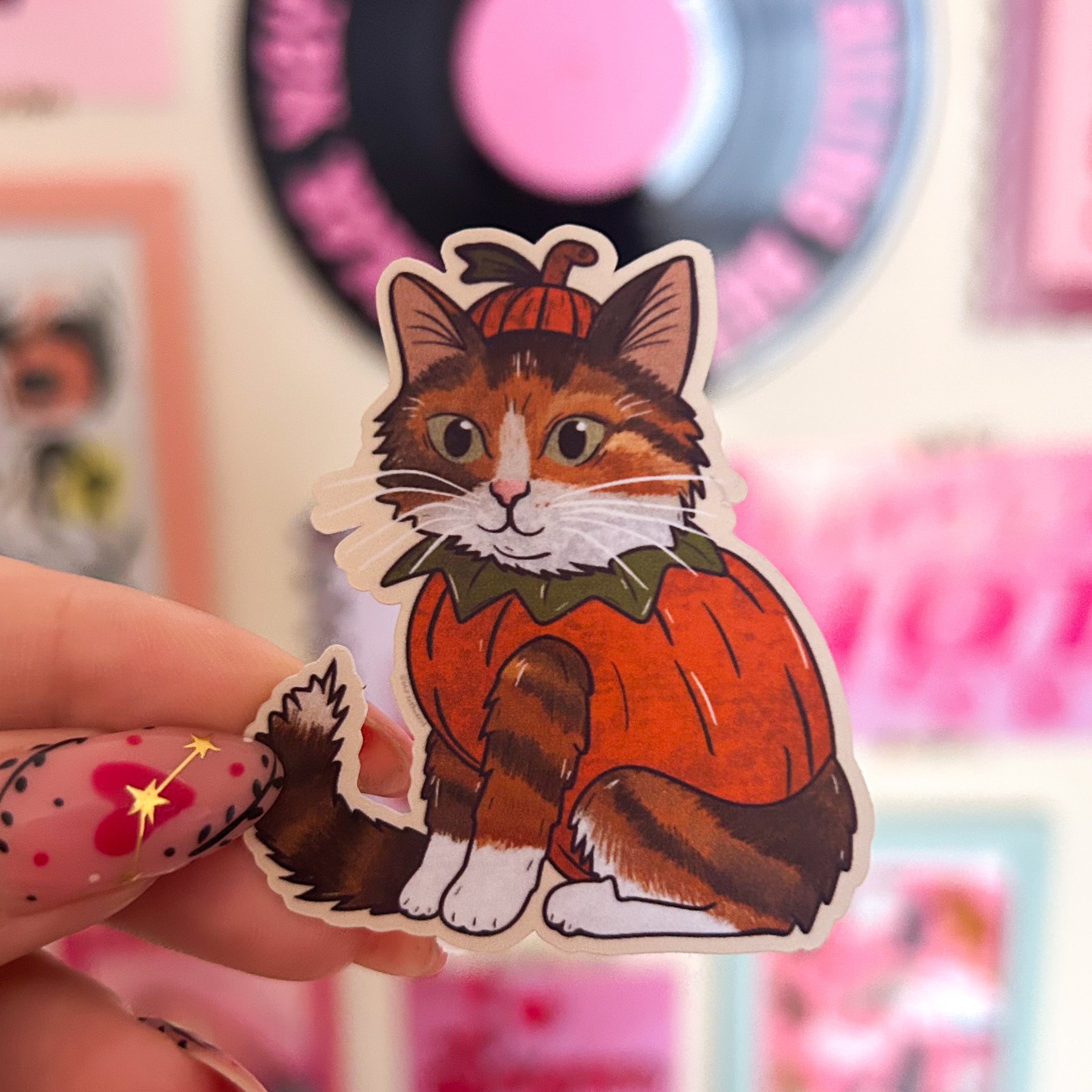 Sticker of a ginger tabby cat in a pumpkin costume held by a hand with colorful nail art.