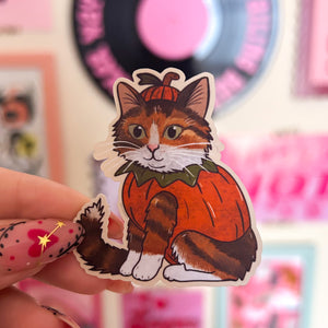 Sticker of a ginger tabby cat in a pumpkin costume held by a hand with colorful nail art.