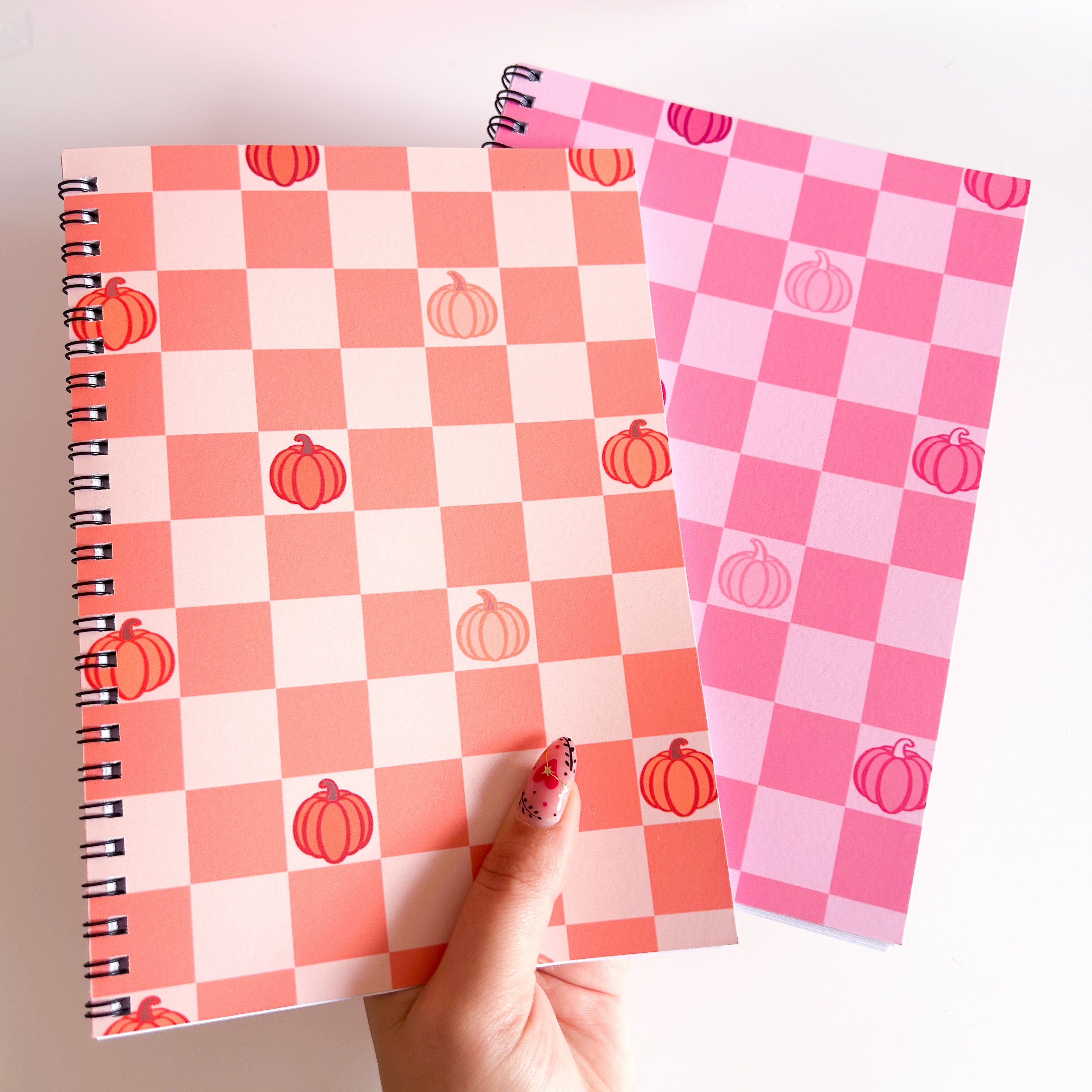 Two checkered notebooks, Orange and Pink, with pumpkin designs held by a hand on a light background