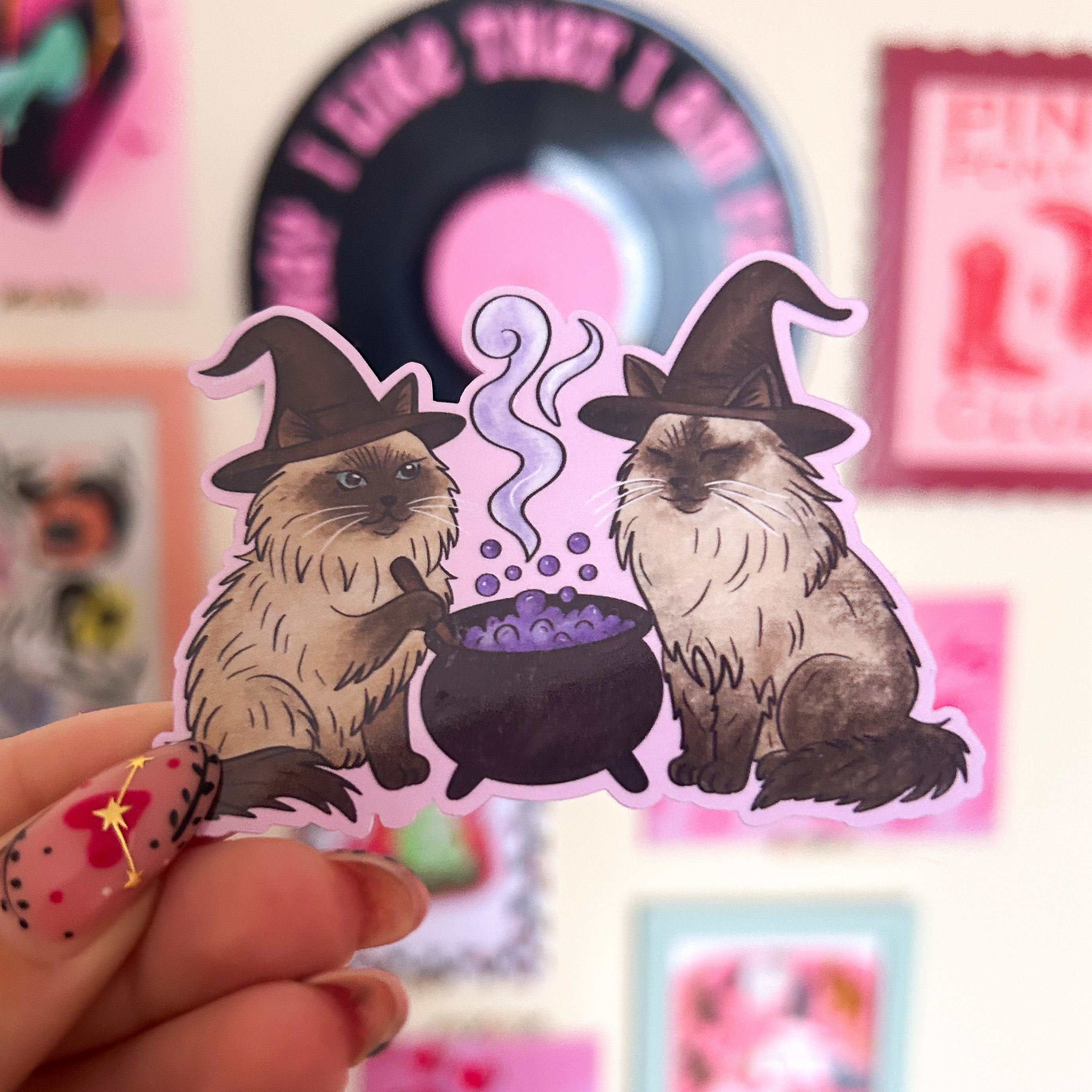 Two ragdoll cats wearing witch hats with a cauldron on a pink background.